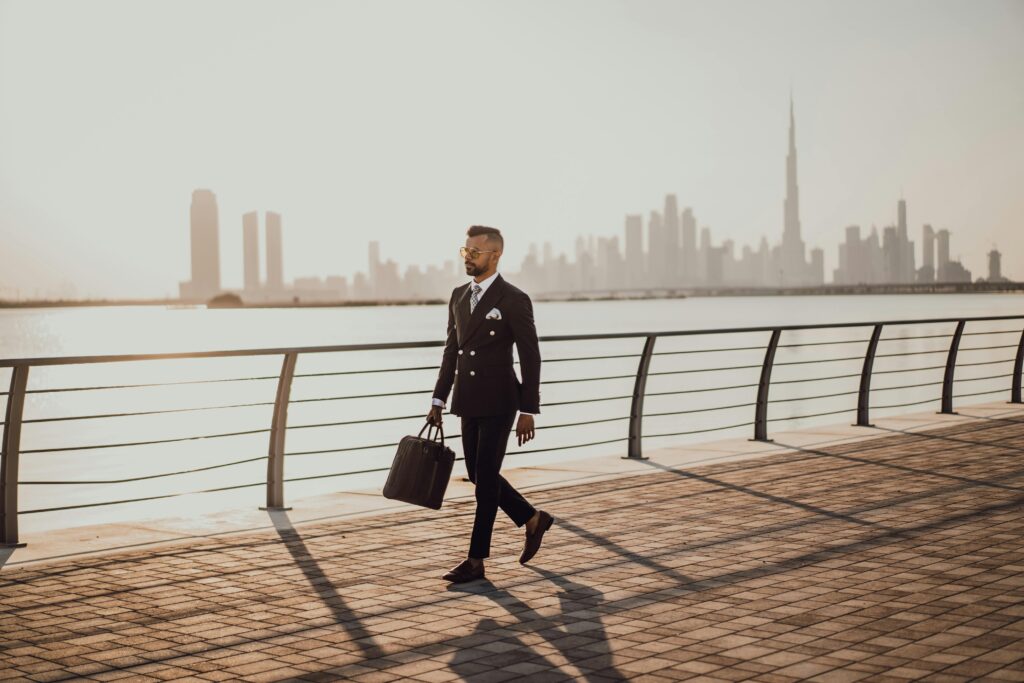 Professional man in formal suit walking by Dubai waterfront with city skyline and Burj Khalifa in view.