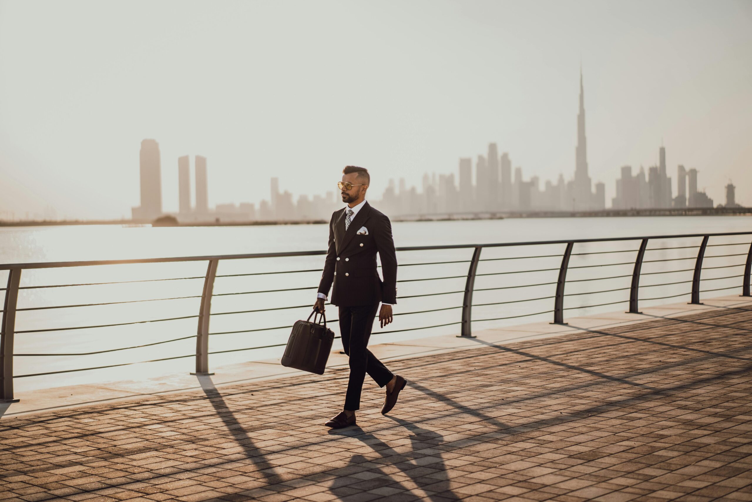 Home Professional man in formal suit walking by Dubai waterfront with city skyline and Burj Khalifa in view.