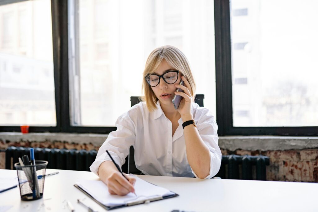 A woman in an office setting multitasking with a phone call and taking notes.